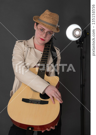 Young female hipster musician holding Spanish six string acoustic guitar in front of her hugging string instrument with both hands and looking at camera. Woman in beige leather jacket and straw hat Young female hipster musician holding Spanish six string acoustic guitar in front of her hugging string instrument with both hands and looking at camera. Woman in beige leather jacket and straw hat 135158556