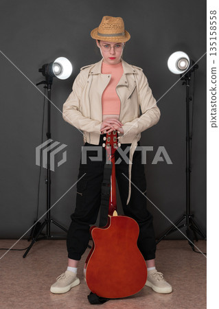 Full length young female guitarist with classical six string acoustic guitar standing on stage and looking at camera. Woman musician is dressed in beige leather jacket, straw hat, black denim joggers Full length young female guitarist with classical six string acoustic guitar standing on stage and looking at camera. Woman musician is dressed in beige leather jacket, straw hat, black denim joggers 135158558
