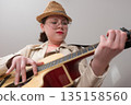 Young female guitarist with glasses and straw hat is practicing classical six string acoustic guitar. Low angle view and dynamic perspective create an exuberant, lively atmosphere, inspiring learning Young female guitarist with glasses and straw hat is practicing classical six string acoustic guitar. Low angle view and dynamic perspective create an exuberant, lively atmosphere, inspiring learning 135158560
