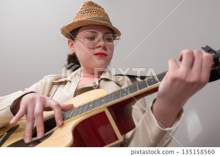 Young female guitarist with glasses and straw hat is practicing classical six string acoustic guitar. Low angle view and dynamic perspective create an exuberant, lively atmosphere, inspiring learning 135158560
