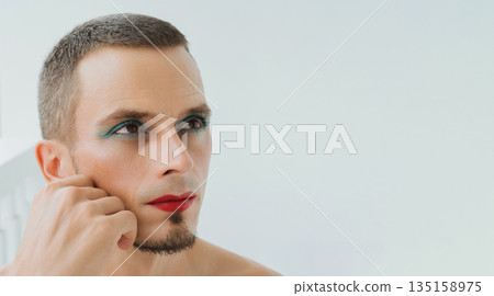 adult gay man with makeup and cosmetics on his face. A handsome transgender male with red lipstick on lips and mascara on eyes on white background adult gay man with makeup and cosmetics on his face. A handsome transgender male with red lipstick on lips and mascara on eyes on white background 135158975