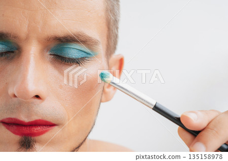 portrait of a feminine gay man with makeup and cosmetics on face. A handsome transgender male with red lipstick applied eye shadow sparkle on his eyes with a brush on white background portrait of a feminine gay man with makeup and cosmetics on face. A handsome transgender male with red lipstick applied eye shadow sparkle on his eyes with a brush on white background 135158978
