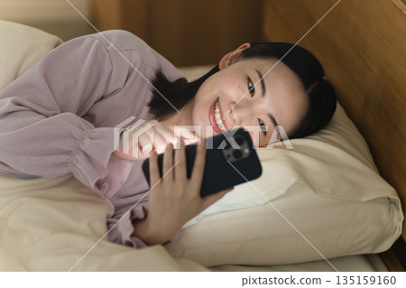 一位年輕女子在床上操作智慧型手機生活風格影像 一位年輕女子在床上操作智慧型手機生活風格影像 135159160