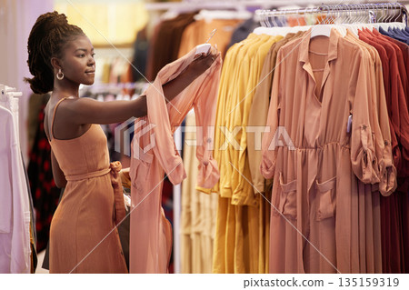 Black woman, shopping and fashion rail with retail choice at a shop with discount and deal. Mall, clothes and African female person at a apparel sale holding a dress with a customer at a boutique Black woman, shopping and fashion rail with retail choice at a shop with discount and deal. Mall, clothes and African female person at a apparel sale holding a dress with a customer at a boutique 135159319