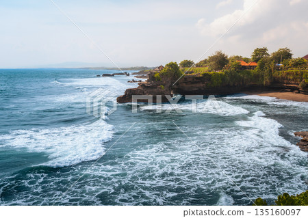 Tanah Lot temple Bali ocean waves crashing rock, iconic Hindu shrine on cliff amid turquoise sea lush greenery blue sky exotic tropical Indonesian coastal paradise serene landscape travel destination 135160097