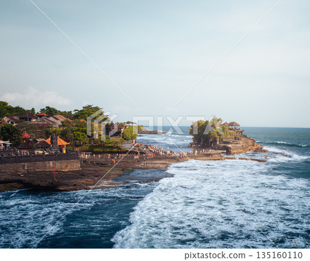 Tanah Lot temple Bali ocean waves crashing rock, iconic Hindu shrine on offshore cliff amid turquoise sea lush greenery orange flags blue sky exotic tropical Indonesian coastal paradise landscape 135160110