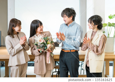 Smiling businesswoman receiving a bouquet of flowers and her team members applauding 135161625