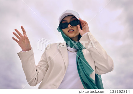 Portrait, fashion or religion with a muslim woman on a cloudy sky background in a cap and scarf for modern style. Islam, faith or hijab with a trendy young arab person outside in contemporary clothes 135162814