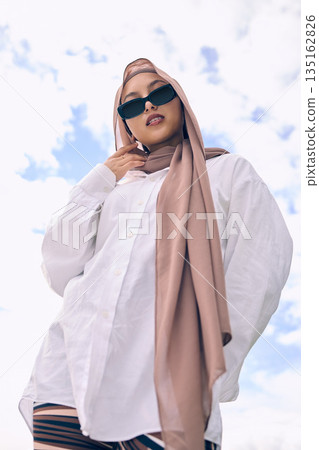 Portrait, fashion and a muslim woman outdoor on a blue sky background in sunglasses and scarf for contemporary style. Islam, faith and hijab with a trendy young arab person outside in modern clothing Portrait, fashion and a muslim woman outdoor on a blue sky background in sunglasses and scarf for contemporary style. Islam, faith and hijab with a trendy young arab person outside in modern clothing 135162826