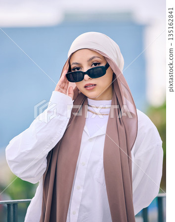Portrait, fashion or sunglasses with a muslim woman in dubai wearing a cap and scarf for contemporary style. Islamic, faith and hijab with a trendy young arab person posing outside in modern eyewear Portrait, fashion or sunglasses with a muslim woman in dubai wearing a cap and scarf for contemporary style. Islamic, faith and hijab with a trendy young arab person posing outside in modern eyewear 135162841