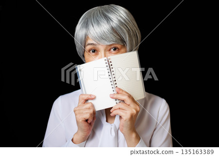A gray-haired senior woman wearing a blouse grinning while hiding half of her face with a notepad 135163389