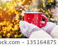 Female hands in warm knitted gloves holding a red mug with a hot drink  against the lights of a Christmas tree in winter 135164875