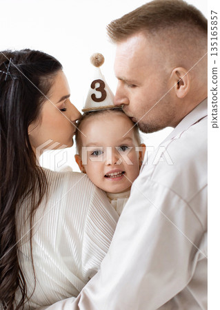A loving family celebrates a child's third birthday with kisses. The parents embrace their daughter, who wears a party hat. A loving family celebrates a child's third birthday with kisses. The parents embrace their daughter, who wears a party hat. 135165857