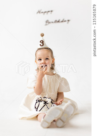A young girl celebrates her third birthday, enjoying a piece of cake while wearing a party hat. A young girl celebrates her third birthday, enjoying a piece of cake while wearing a party hat. 135165879