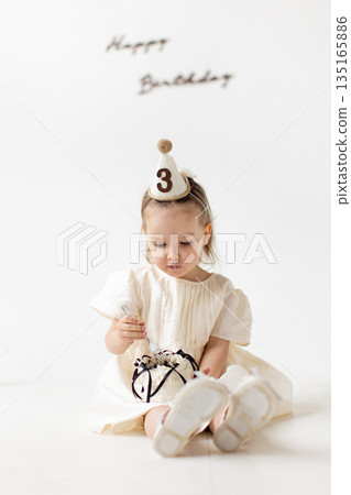 A young girl celebrates her third birthday, sitting and enjoying a cake with a party hat on. The background is plain white. A young girl celebrates her third birthday, sitting and enjoying a cake with a party hat on. The background is plain white. 135165886