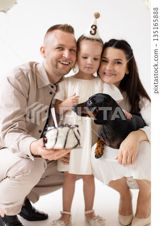 A family celebrates a child's third birthday with a cake and a dachshund dog in a bright, white setting. A family celebrates a child's third birthday with a cake and a dachshund dog in a bright, white setting. 135165888