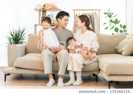 一家人笑容滿面地抱著嬰兒和女兒坐在家裡的沙發上,照片中是父母和孩子。 一家人笑容滿面地抱著嬰兒和女兒坐在家裡的沙發上,照片中是父母和孩子。 135166159
