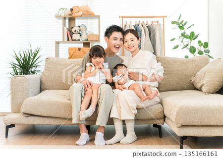 一家人笑容滿面地抱著嬰兒和女兒坐在家裡的沙發上，照片中是父母和孩子。 135166161