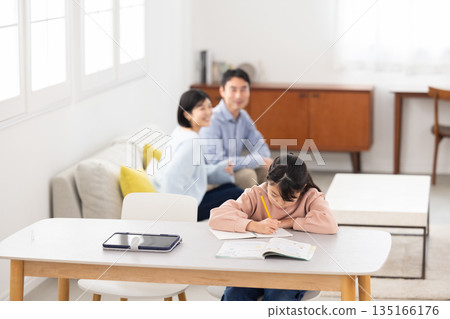 An elementary school girl studying at the living room table while her parents watch An elementary school girl studying at the living room table while her parents watch 135166176