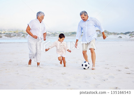 Beach, football and grandparents playing with kid on vacation or holiday happy for sand soccer or sports. Travel, summer and elderly man and woman kicking a ball with child at the ocean together 135166543