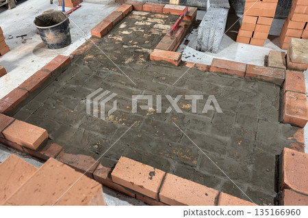 Fresh brickwork of a heating stove with a bench base. Laid out of brick on a concrete screed. Bricks and tools nearby. Fresh brickwork of a heating stove with a bench base. Laid out of brick on a concrete screed. Bricks and tools nearby. 135166960
