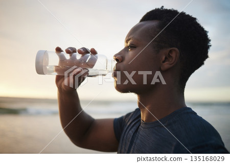 Black man, beach fitness and drinking water at sunset on running break, exercise or workout. Drink, liquid and African male athlete with bottle for nutrition, hydration or health, wellness and ocean. 135168029
