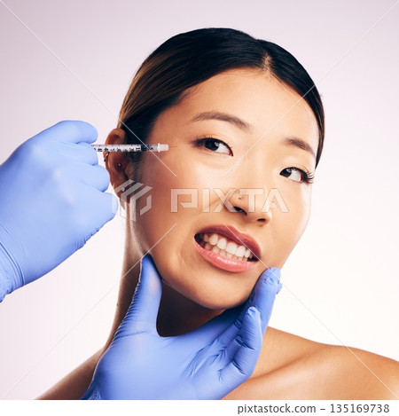 Face injection, skincare and Asian woman in pain in studio isolated on a white background. Cosmetics, syringe and female model with collagen filler, dermatology and prp facelift in plastic surgery. 135169738