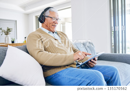 Old man laughing, relax with tablet and headphones at home, retirement and technology, streaming comedy online. Surfing internet, watching funny video and elderly male person sitting on couch 135170562