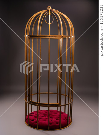 Human-sized golden birdcage with an open door and red velvet cushion on a gray background. Human-sized golden birdcage with an open door and red velvet cushion on a gray background. 135172233