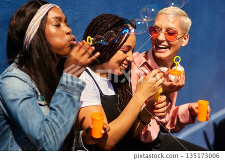Women, friends, and blowing bubbles on summer vacation, holiday travel or bonding activity and against a blue wall. Happy, game and ladies playing with soap or wand toy or in freedom and social break 135173006