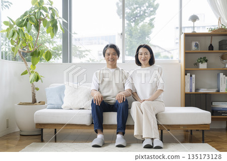 Image of a couple in their 50s sitting side by side on the sofa and smiling at the camera 135173128
