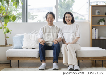 Image of a couple in their 50s sitting side by side on the sofa and smiling at the camera 135173177
