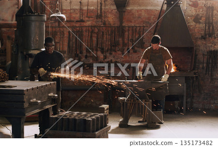 Hammer, anvil and sparks with men working in a forge for metal work manufacturing or production. Industry, welding and trade with a young male blacksmith in a workshop, plant or industrial foundry Hammer, anvil and sparks with men working in a forge for metal work manufacturing or production. Industry, welding and trade with a young male blacksmith in a workshop, plant or industrial foundry 135173482