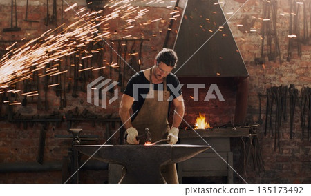 Hammer, anvil and fire with a man working in a plant for metal work manufacturing or production. Industry, welding and trade with a male blacksmith at work in a factory, forge or industrial workshop Hammer, anvil and fire with a man working in a plant for metal work manufacturing or production. Industry, welding and trade with a male blacksmith at work in a factory, forge or industrial workshop 135173492