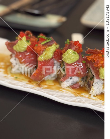 Close-up of colorful tuna sushi roll topped with roe, wasabi, and microgreens, served on a rectangular ceramic dish. Close-up of colorful tuna sushi roll topped with roe, wasabi, and microgreens, served on a rectangular ceramic dish. 135173942