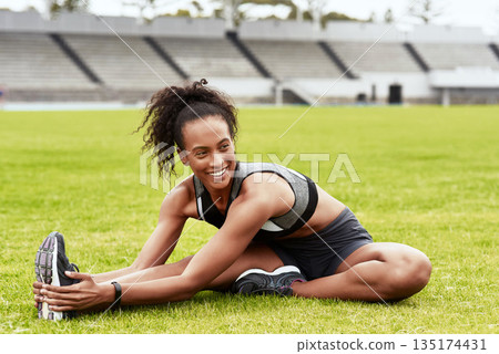 Happy woman, fitness and stretching body on grass for running, exercise or workout at the stadium. Active female person or athlete in warm up leg stretch for sports training or cardio run on a field 135174431