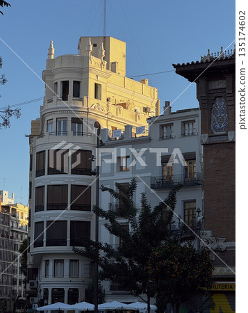 Elegant white corner building with rounded edges and rooftop tower, lit by evening sun in central Valencia. 135174602