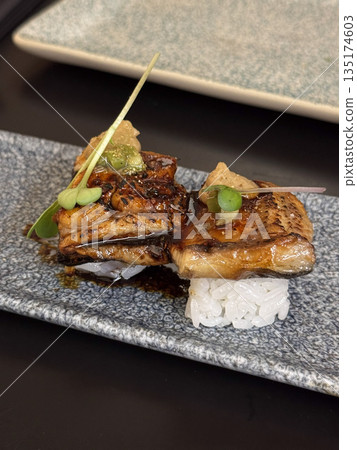 Elegant Japanese dish of unagi nigiri sushi garnished with herbs and sauce, served on a modern slate plate. Elegant Japanese dish of unagi nigiri sushi garnished with herbs and sauce, served on a modern slate plate. 135174603