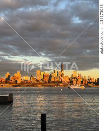 Manhattan skyline lit by golden hour sunlight, with dramatic clouds over water and reflections at sunset. Manhattan skyline lit by golden hour sunlight, with dramatic clouds over water and reflections at sunset. 135175088