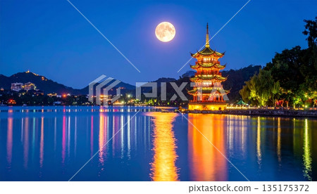 The nighttime scene is reflected on the lake's surface under the dark sky, featuring striking traditional architecture and tall pagodas. 135175372