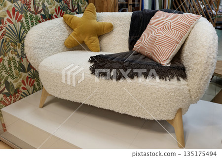 A white cozy sofa is on display with a yellow star pillow and black throw blanket. The backdrop shows colorful patterned wallpaper in a retail space A white cozy sofa is on display with a yellow star pillow and black throw blanket. The backdrop shows colorful patterned wallpaper in a retail space 135175494