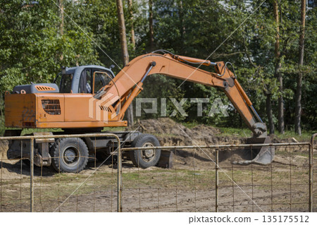 A large excavator digs into the ground at a construction site surrounded by trees during day. Workers may be nearby managing the project 135175512