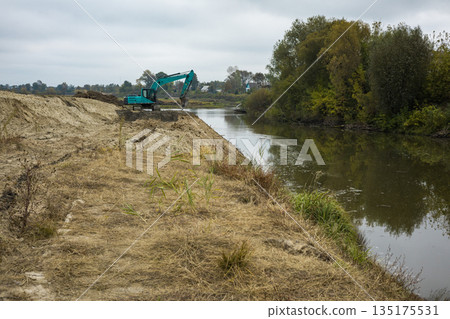 A machine is digging near the riverbank while trees and bushes line the opposite shore. The sky is overcast and the water is still 135175531