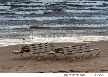 Empty beach chairs on Jurmala shore with waves crashing under cloudy sky 135175569
