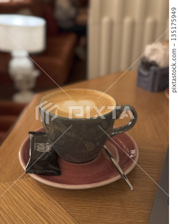 Fresh cappuccino in a ceramic cup served with a saucer and spoon, placed on a cozy wooden cafe table. Fresh cappuccino in a ceramic cup served with a saucer and spoon, placed on a cozy wooden cafe table. 135175949