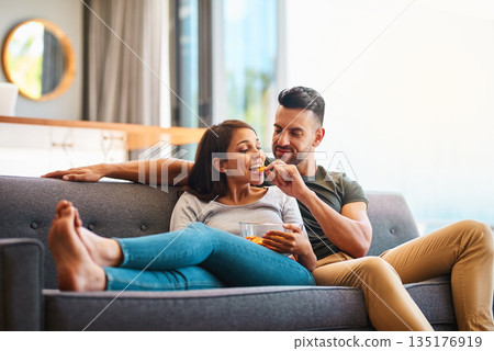Love, happy couple on sofa and eating chips in living room of their home. Caring or bonding together, relaxing or comfortable and happy married people on couch watching television for quality time. Love, happy couple on sofa and eating chips in living room of their home. Caring or bonding together, relaxing or comfortable and happy married people on couch watching television for quality time. 135176919