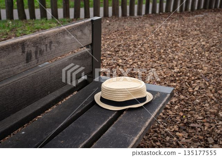 Straw summer hat resting on a wooden park bench outdoors. Simple everyday scene showing a casual personal accessory placed in a public green space. Straw summer hat resting on a wooden park bench outdoors. Simple everyday scene showing a casual personal accessory placed in a public green space. 135177555