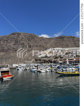 Brightly colored fishing boats floating in a small harbor surrounded by rocky hills and sunny skies in Tenerife. Brightly colored fishing boats floating in a small harbor surrounded by rocky hills and sunny skies in Tenerife. 135177798