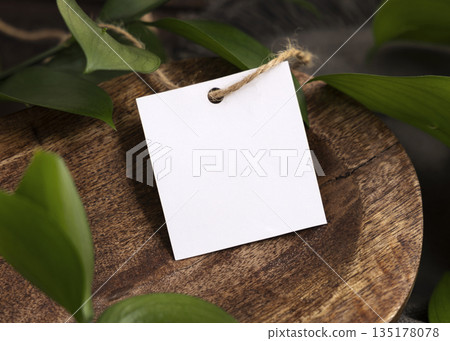Square gift tag mockup on brown wooden tray near green leaves and grey fabric closeup 135178078
