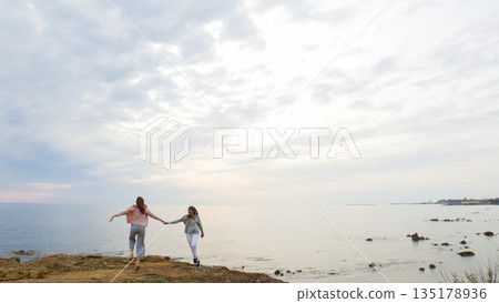 Two young women walking on the rocky shore near the sea and hanging hands 135178936